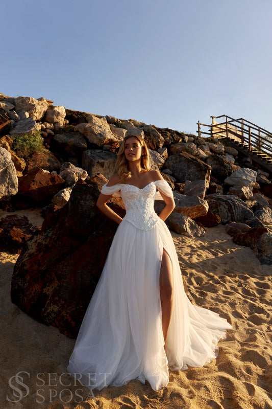 Bride in romantic A-line gown with off-the-shoulder draped tulle sleeves, lace corset bodice, V-neckline, and slit tulle skirt.