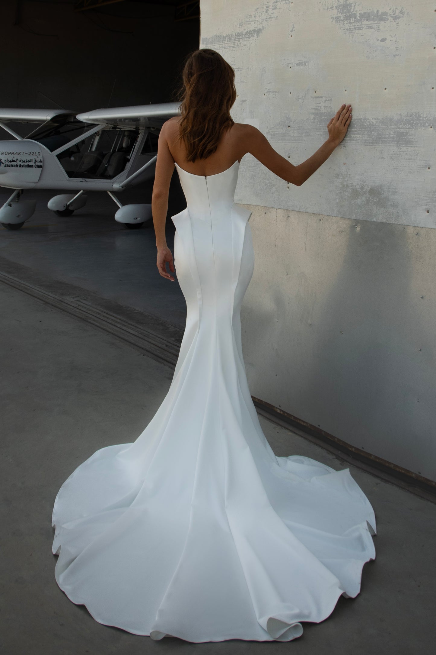 Bride wearing strapless satin mermaid wedding dress with structured sides and chapel train, photographed in a hangar beside an airplane.