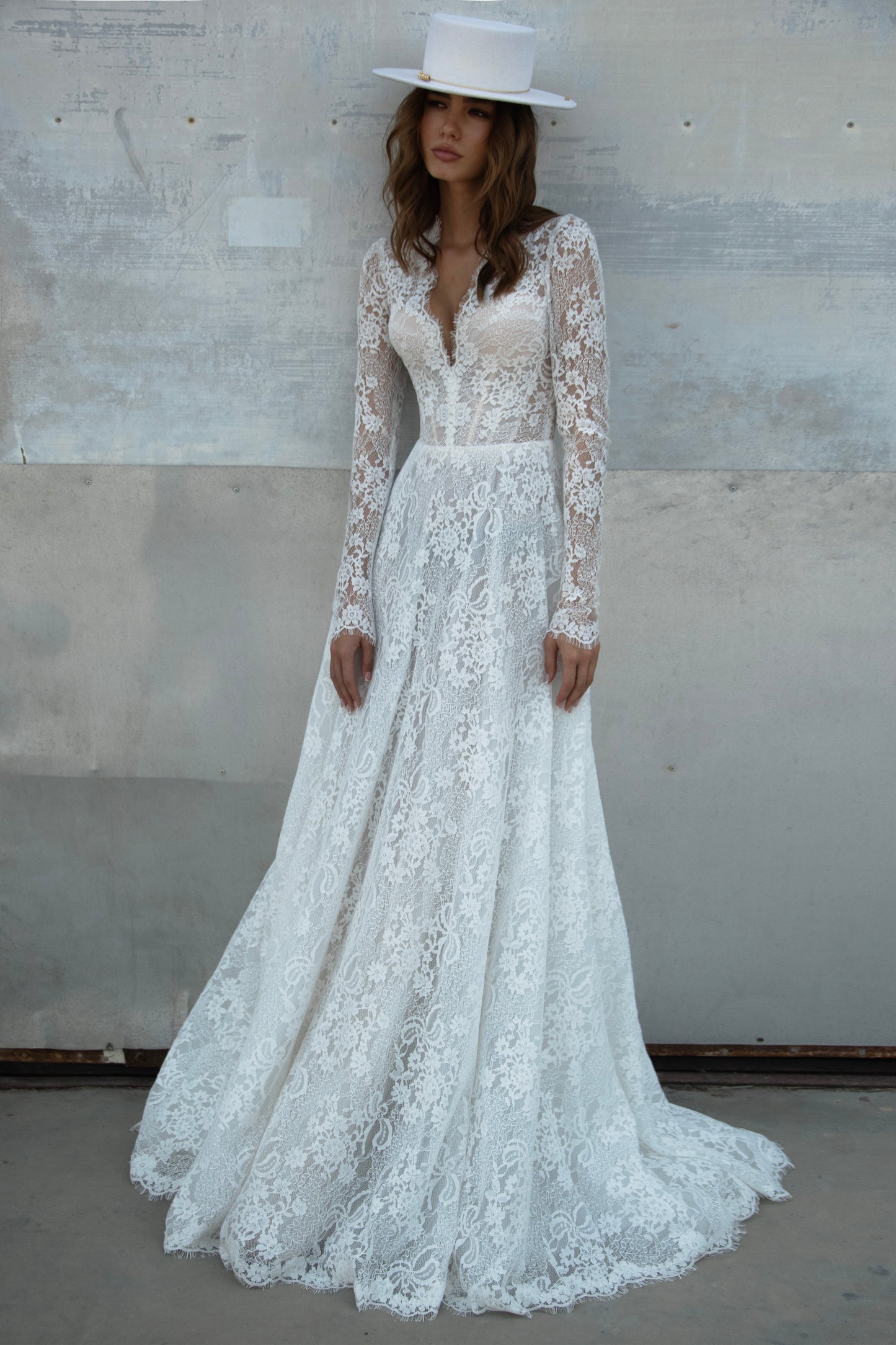 A-line lace wedding dress with long sleeves, V-neckline, high buttoned back, and flowing court-length train featuring detailed lace texture and modern bridal design