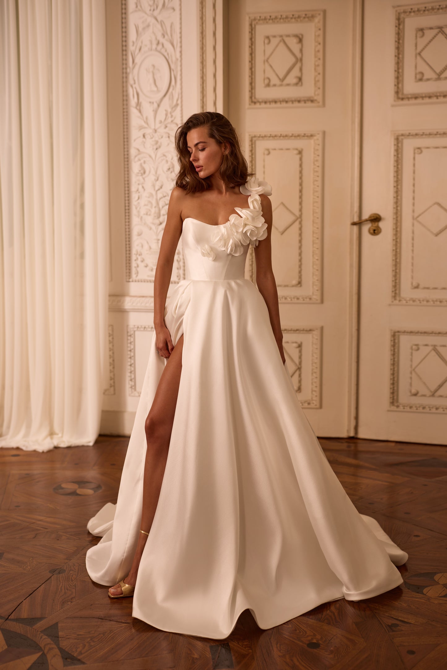 Bride in Mikado A-line wedding dress with one-shoulder floral strap, high slit, and lace-up back, standing confidently in soft natural light.