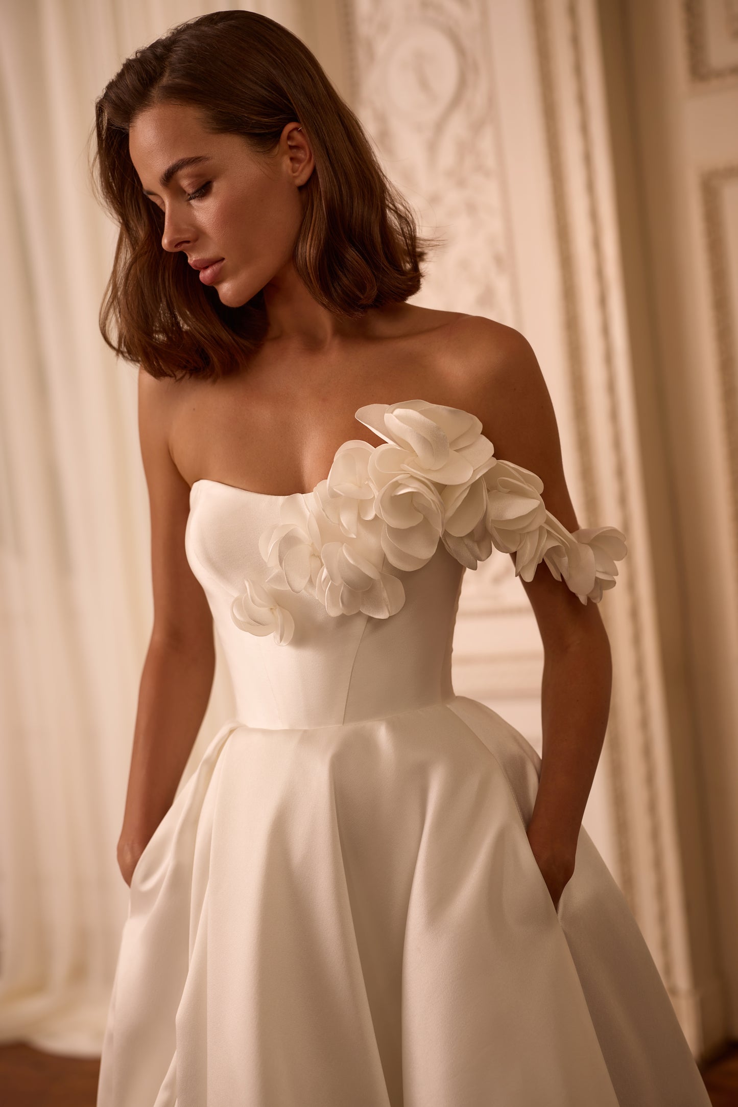 Bride in Mikado A-line wedding dress with one-shoulder floral strap, high slit, and lace-up back, standing confidently in soft natural light.