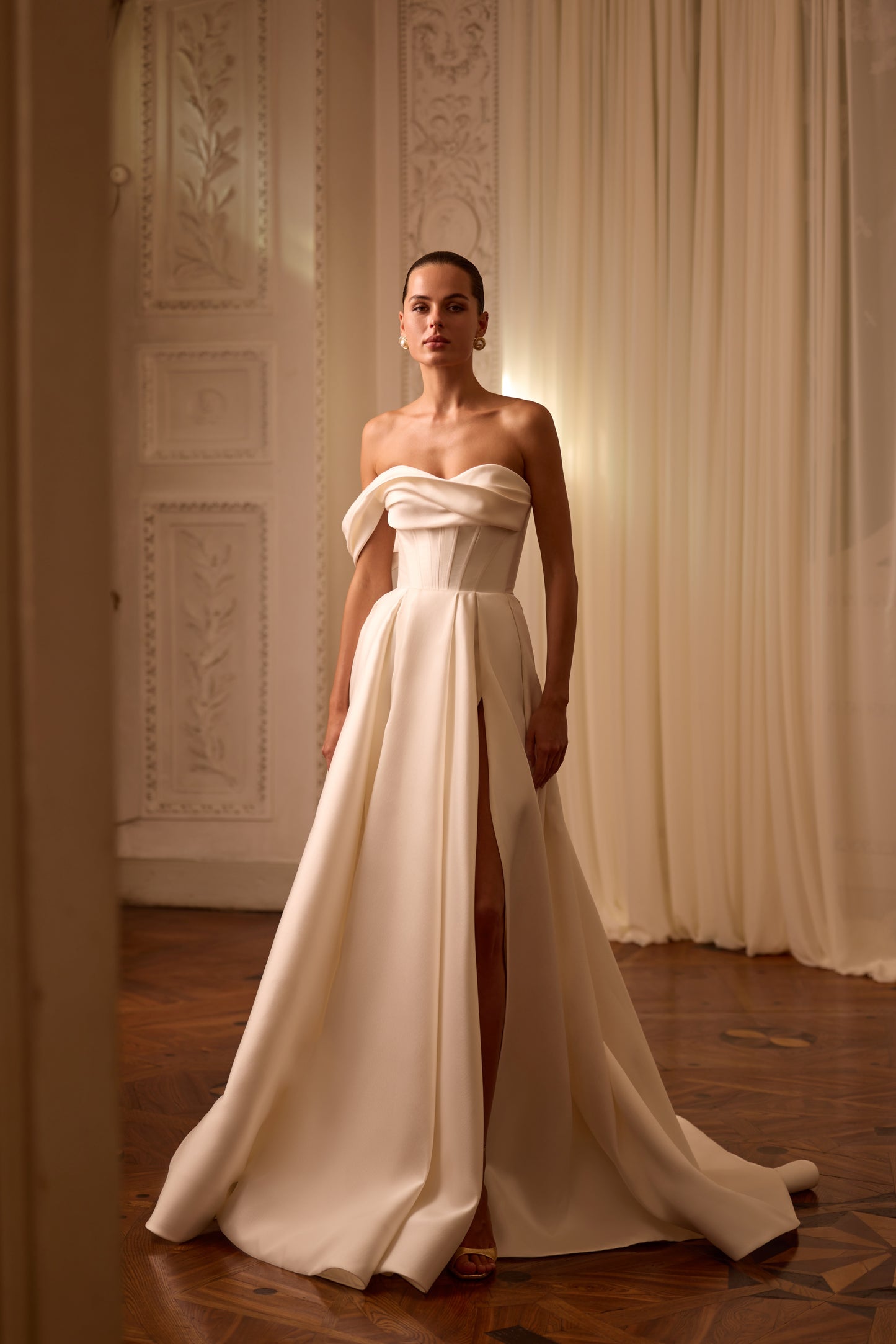 Bride in sleeveless A-line Mikado wedding gown with draped one-shoulder neckline, thigh-high slit, lace-up back, and court train in soft light.