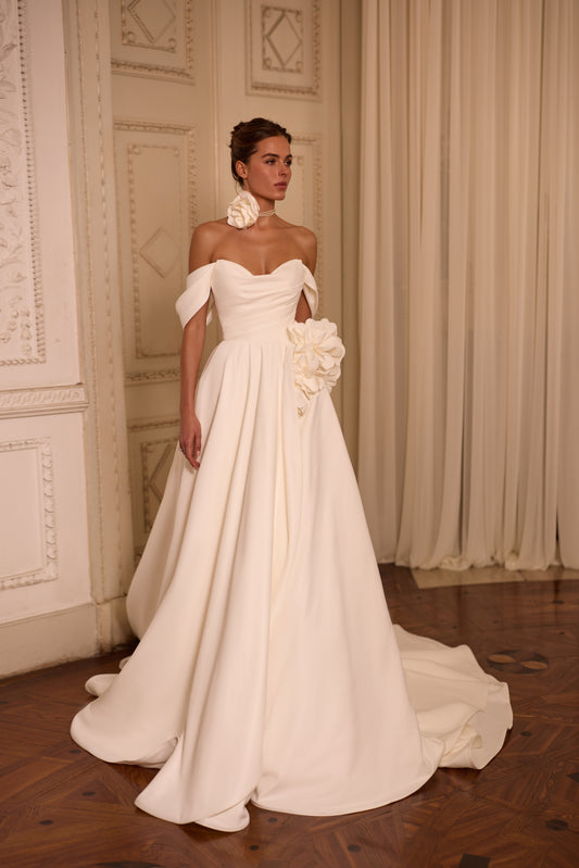 Bride in classic off-the-shoulder A-line wedding gown with draped neckline, lace-up back, floral side detail on skirt, and flowing soft crepe court train.