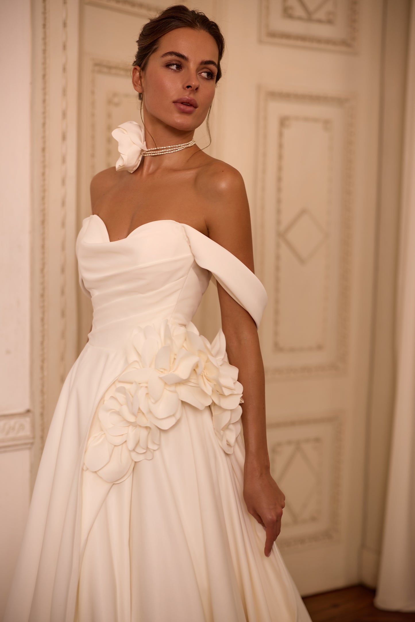 Bride in classic off-the-shoulder A-line wedding gown with draped neckline, lace-up back, floral side detail on skirt, and flowing soft crepe court train.