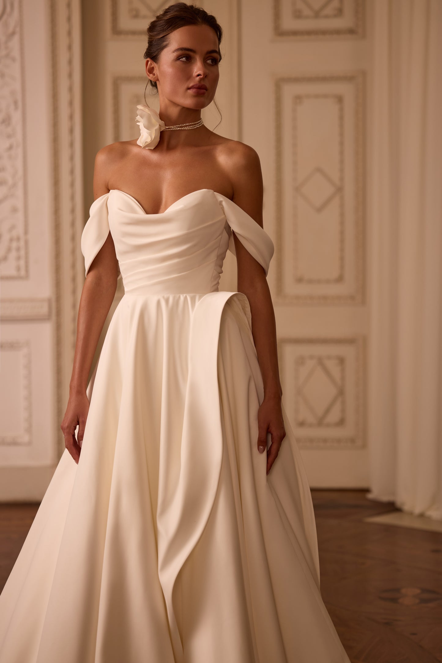 Bride in classic off-the-shoulder A-line wedding gown with draped neckline, lace-up back, floral side detail on skirt, and flowing soft crepe court train.