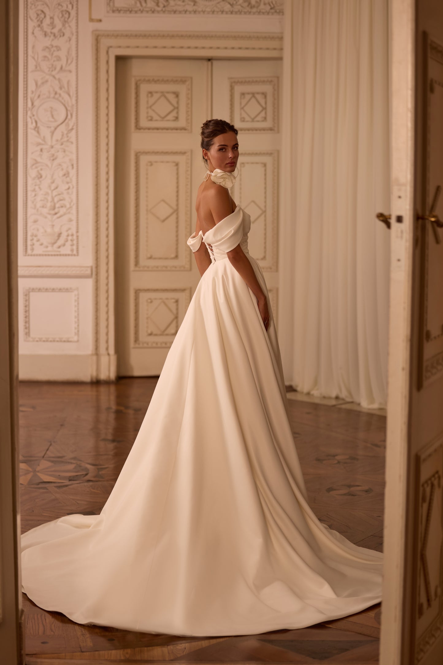 Bride in classic off-the-shoulder A-line wedding gown with draped neckline, lace-up back, floral side detail on skirt, and flowing soft crepe court train.