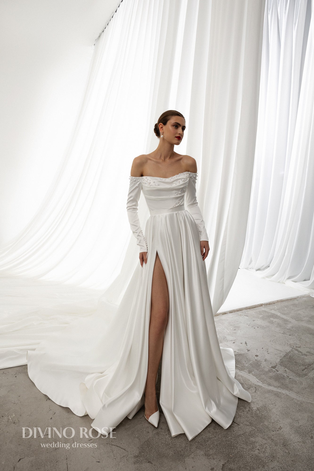 Off-shoulder satin wedding dress with long sleeves and slit.