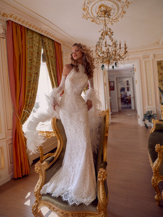 An elegant halter column wedding gown crafted in hand-beaded lace with detachable lace sleeves and a dramatic feather stole. Modern, sculpted, and exquisitely glamorous.