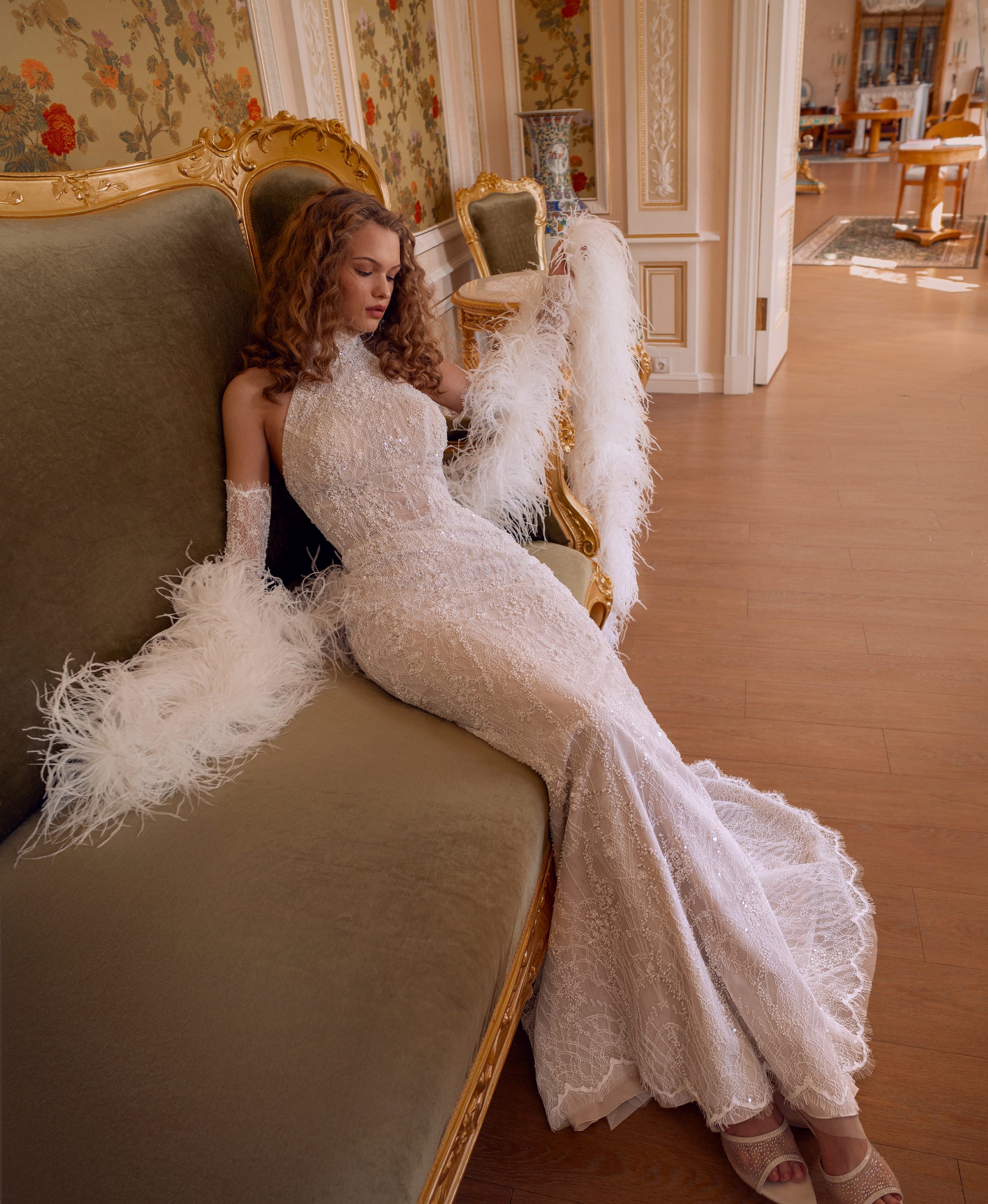 An elegant halter column wedding gown crafted in hand-beaded lace with detachable lace sleeves and a dramatic feather stole. Modern, sculpted, and exquisitely glamorous.