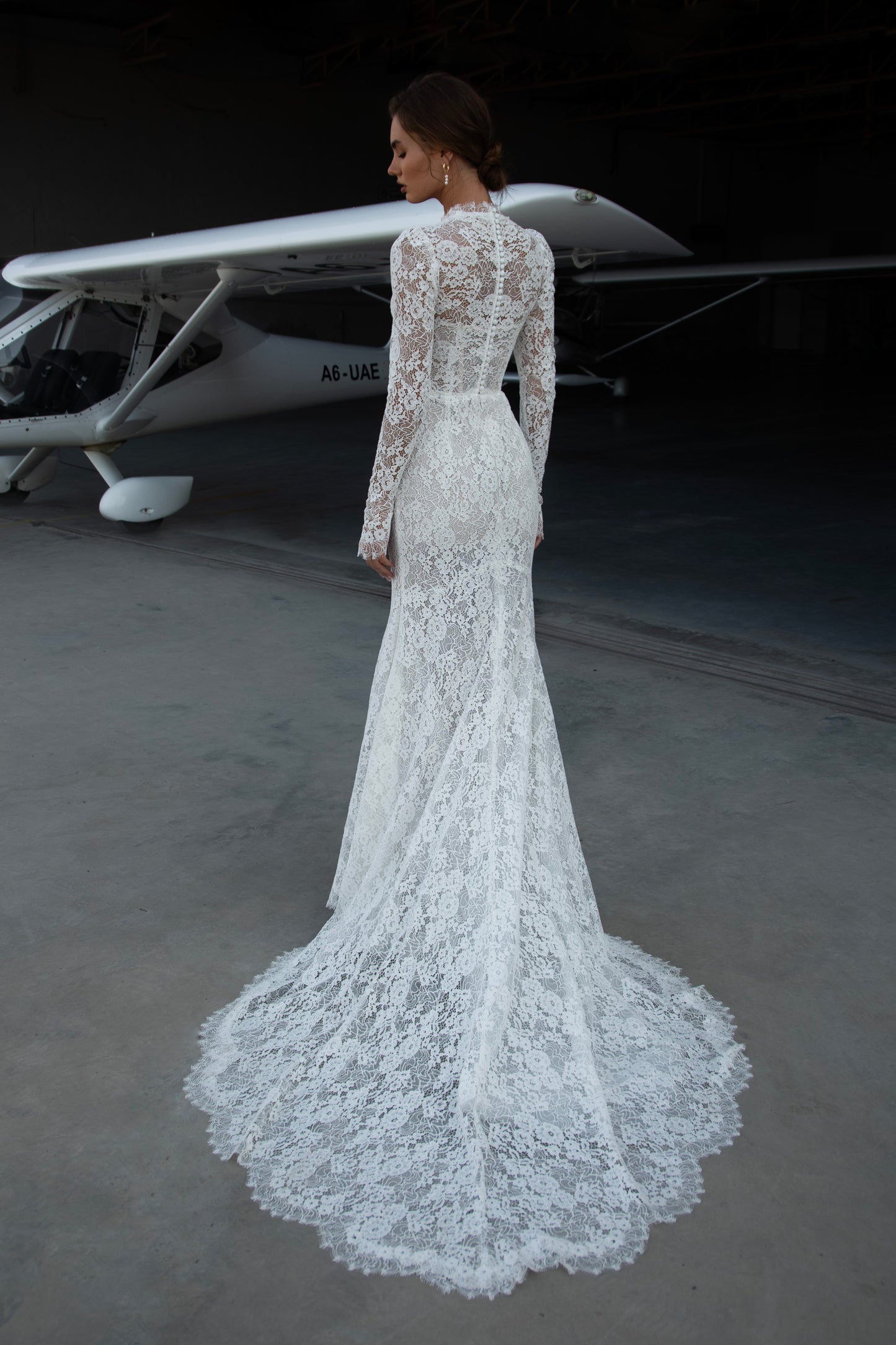 Bride wearing long-sleeve lace fit-and-flare wedding dress with scalloped neckline and chapel train, photographed beside an airplane hangar.