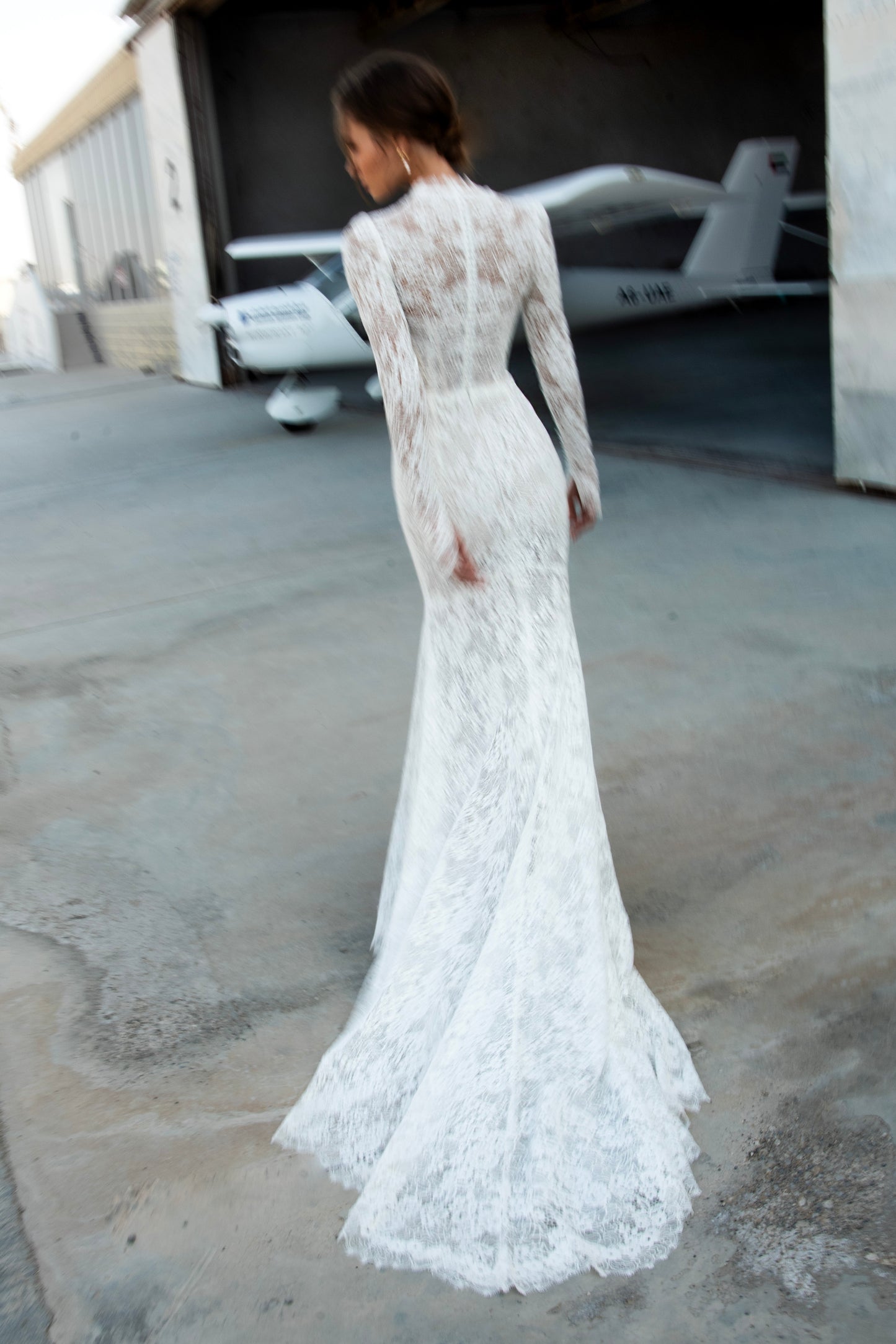 Bride wearing long-sleeve lace fit-and-flare wedding dress with scalloped neckline and chapel train, photographed beside an airplane hangar.