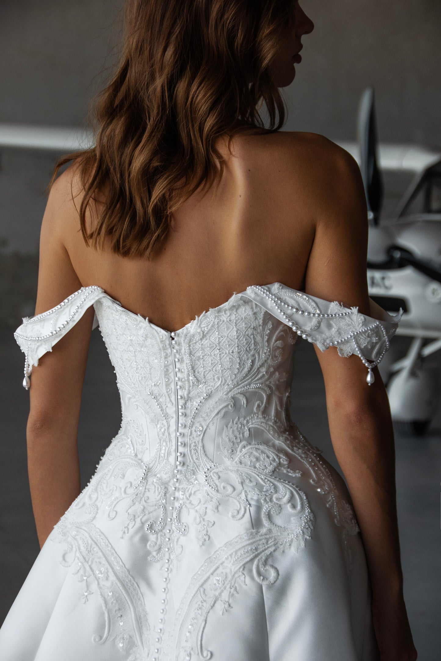 Bride wearing off-the-shoulder Mikado A-line wedding gown with beaded lace bodice and flowing skirt, photographed at an airfield hangar.
