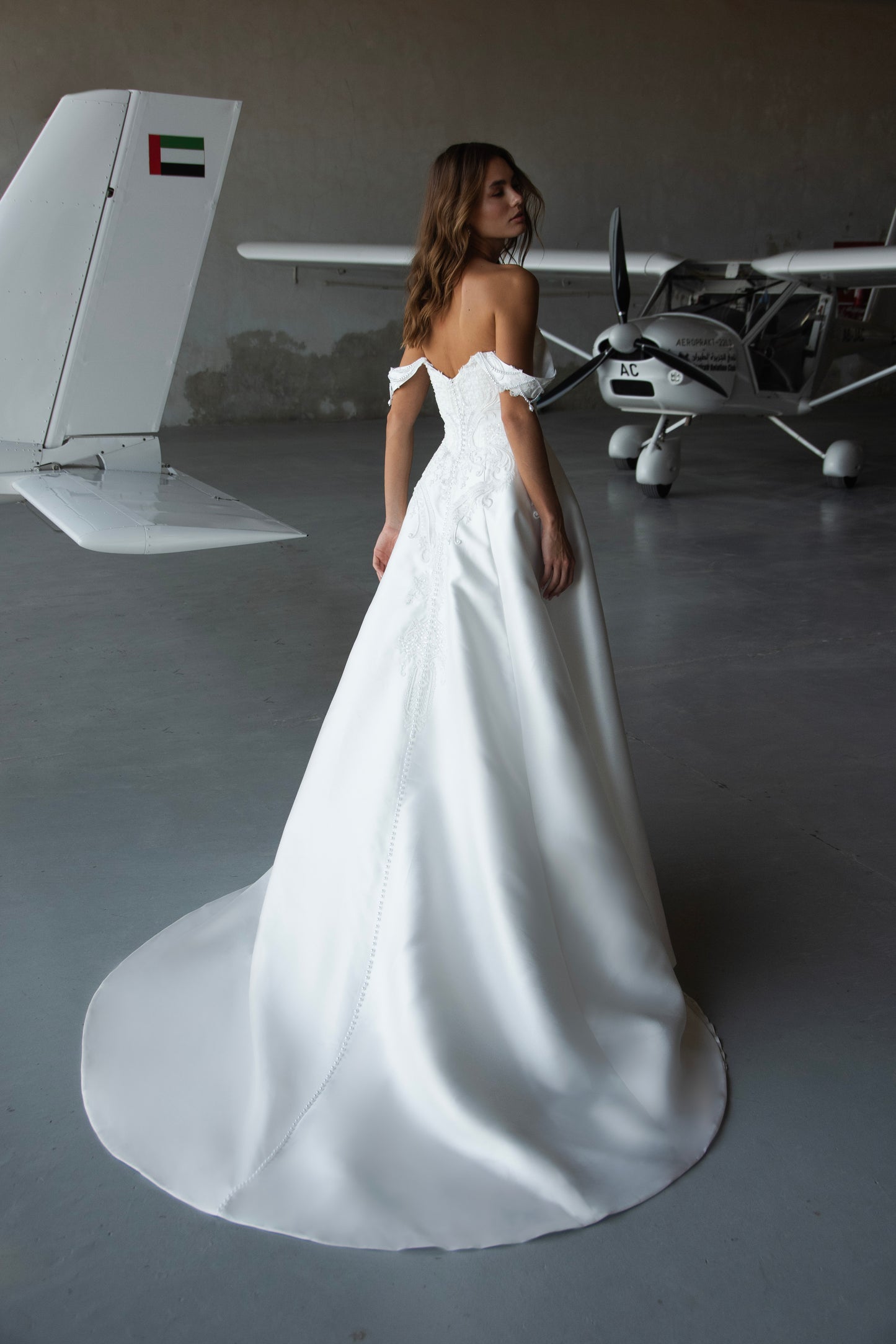 Bride wearing off-the-shoulder Mikado A-line wedding gown with beaded lace bodice and flowing skirt, photographed at an airfield hangar.