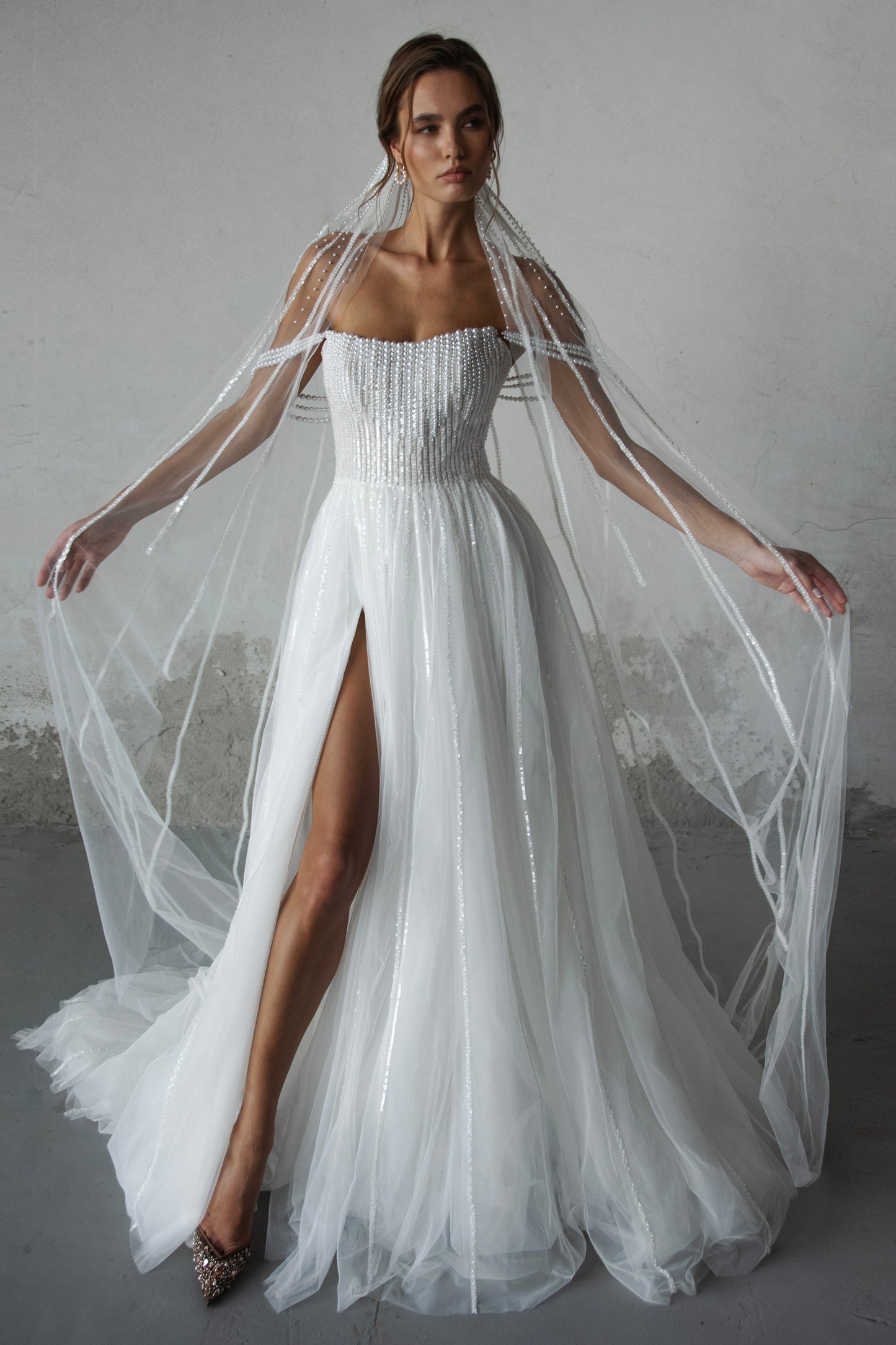 Pearl-embroidered tulle wedding dress with beaded corset bodice, detachable sheer sleeves, thigh-high slit, and flowing layered skirt