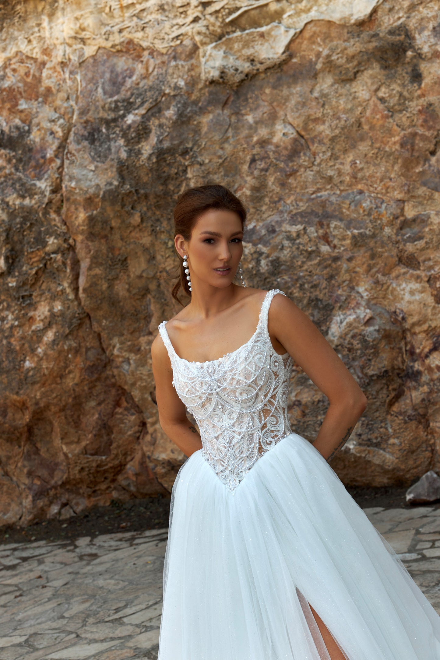 Bride wearing sleeveless A-line wedding gown with boat neckline, pearl and sequin lace bodice, glitter tulle slit skirt, open scoop back, and zip closure.