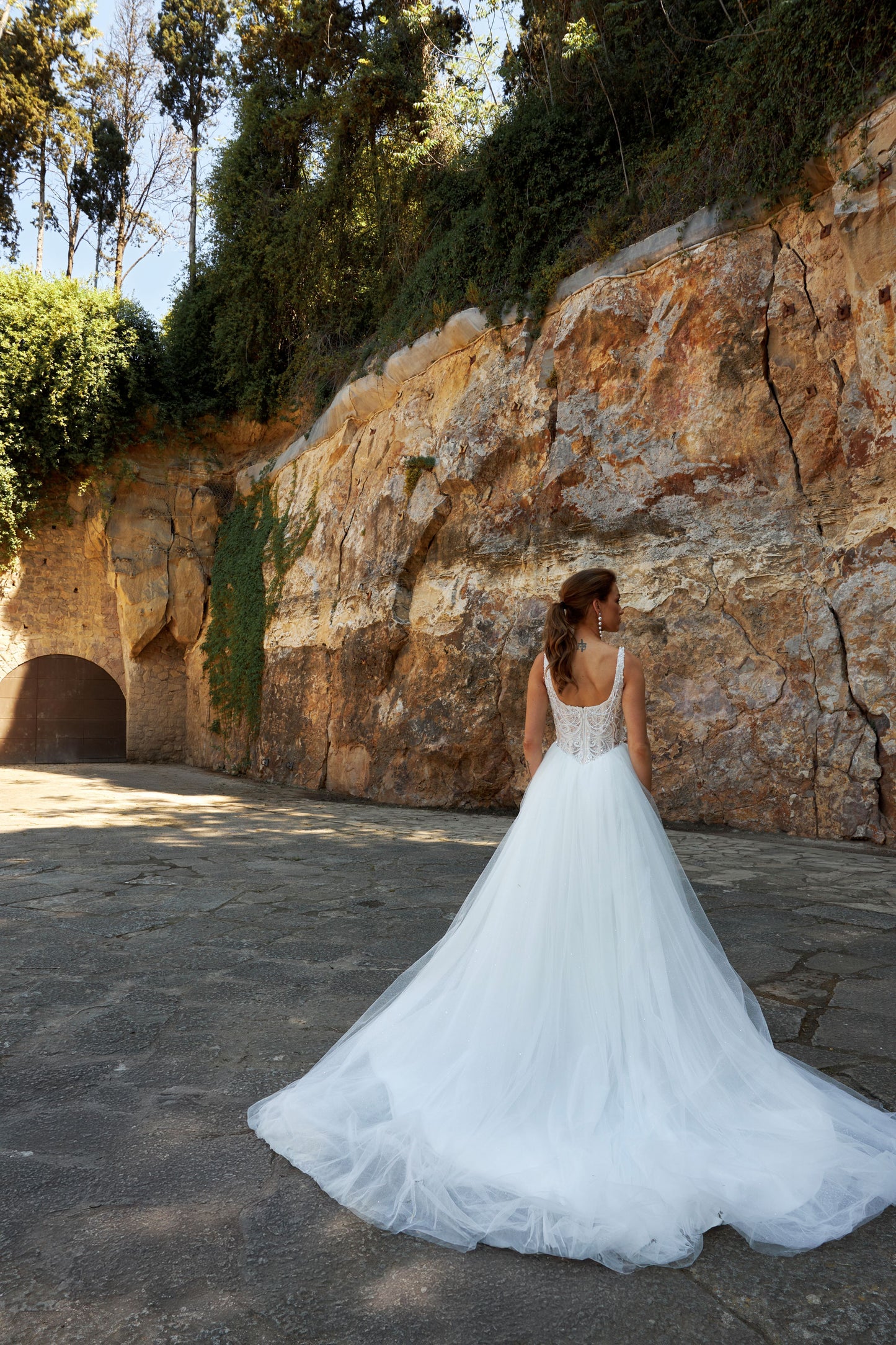 Bride wearing sleeveless A-line wedding gown with boat neckline, pearl and sequin lace bodice, glitter tulle slit skirt, open scoop back, and zip closure.