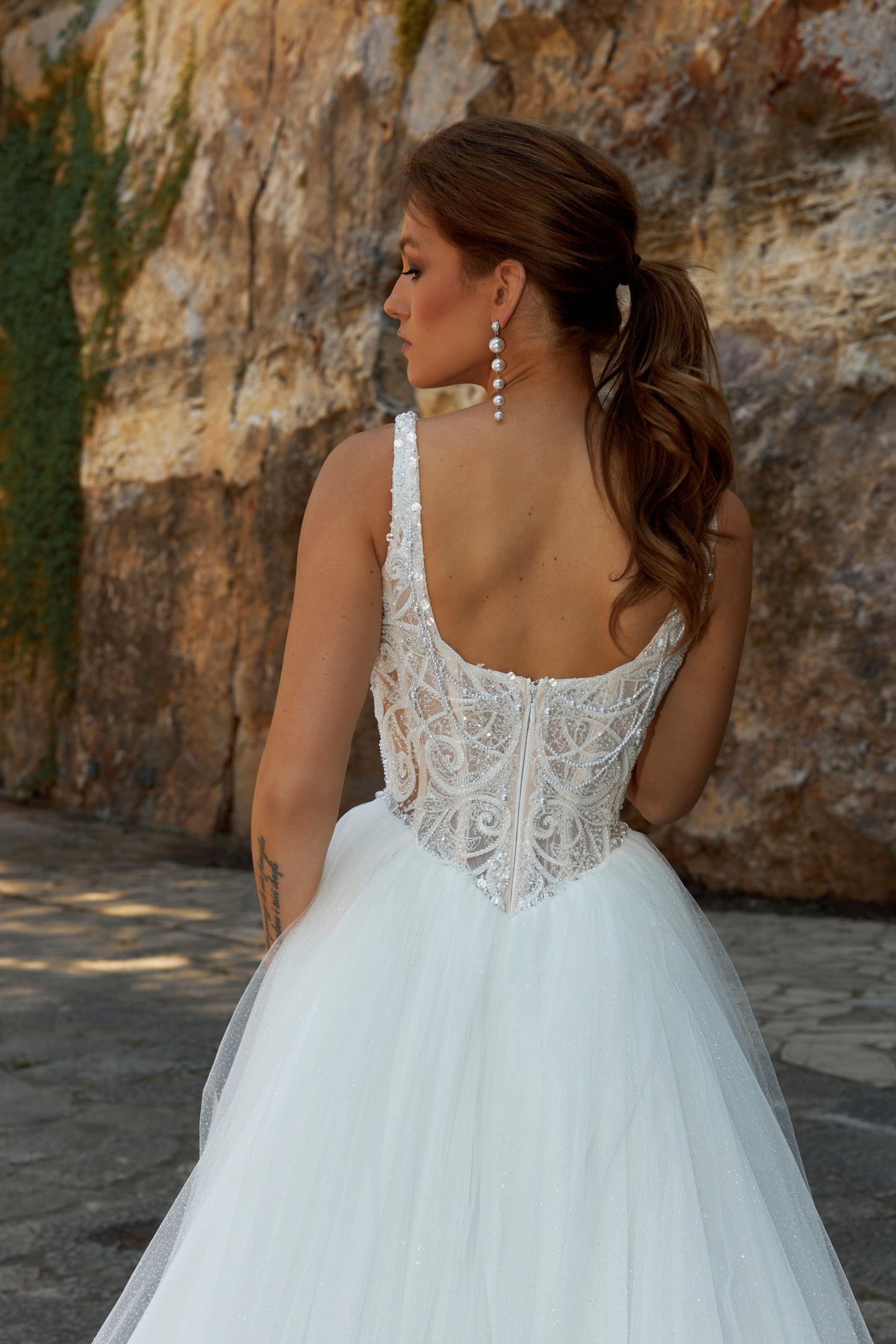 Bride wearing sleeveless A-line wedding gown with boat neckline, pearl and sequin lace bodice, glitter tulle slit skirt, open scoop back, and zip closure.