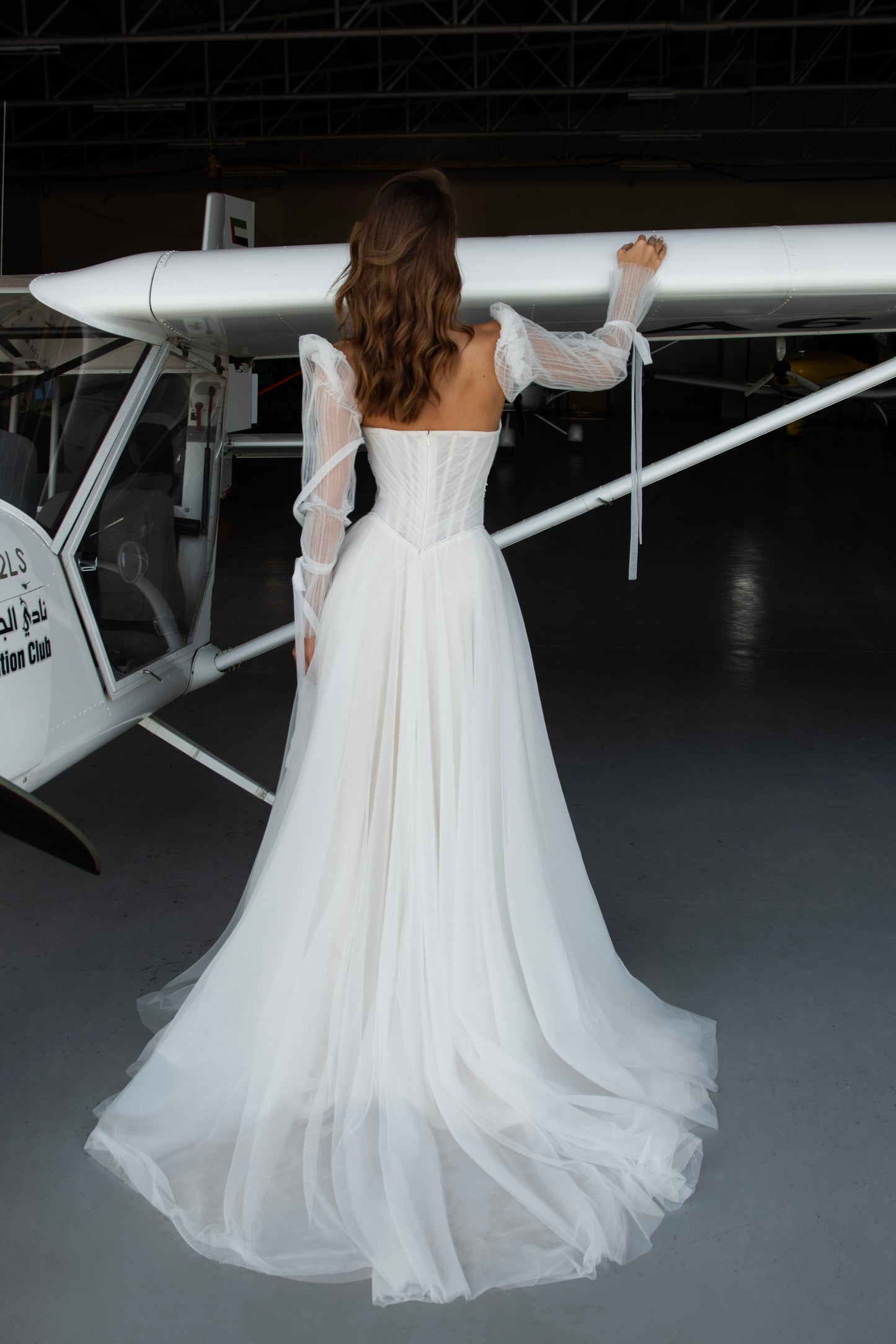 Bride in tulle A-line wedding dress with puff sleeves, pearl-draped corset bodice, and front slit, standing beside an airplane.