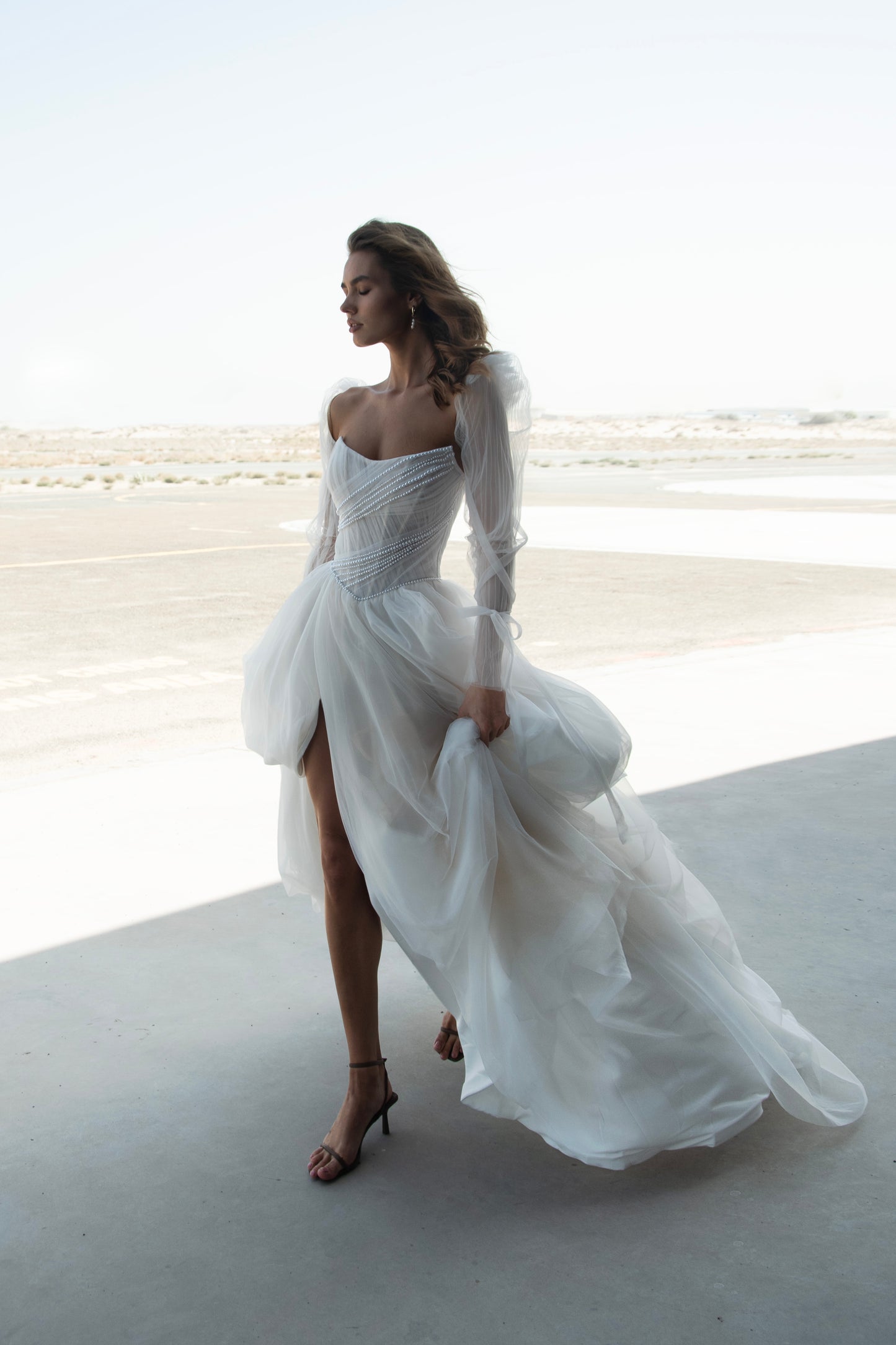 Bride in tulle A-line wedding dress with puff sleeves, pearl-draped corset bodice, and front slit, standing beside an airplane.