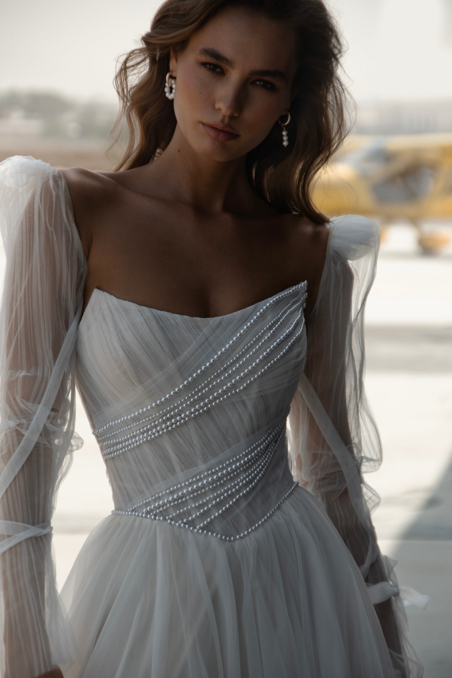 Bride in tulle A-line wedding dress with puff sleeves, pearl-draped corset bodice, and front slit, standing beside an airplane.