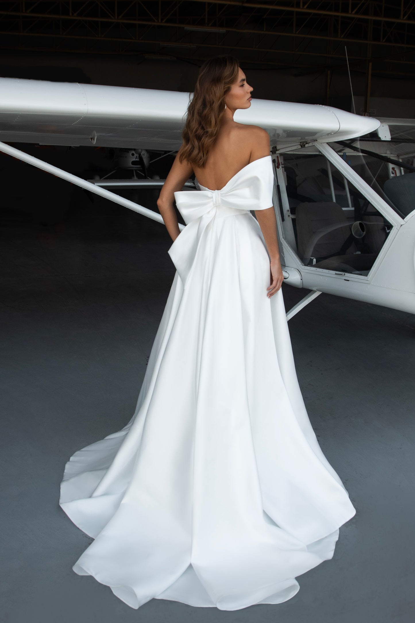 Bride wearing Mikado A-line off-shoulder wedding dress with draped neckline and front slit, standing beside an airplane.