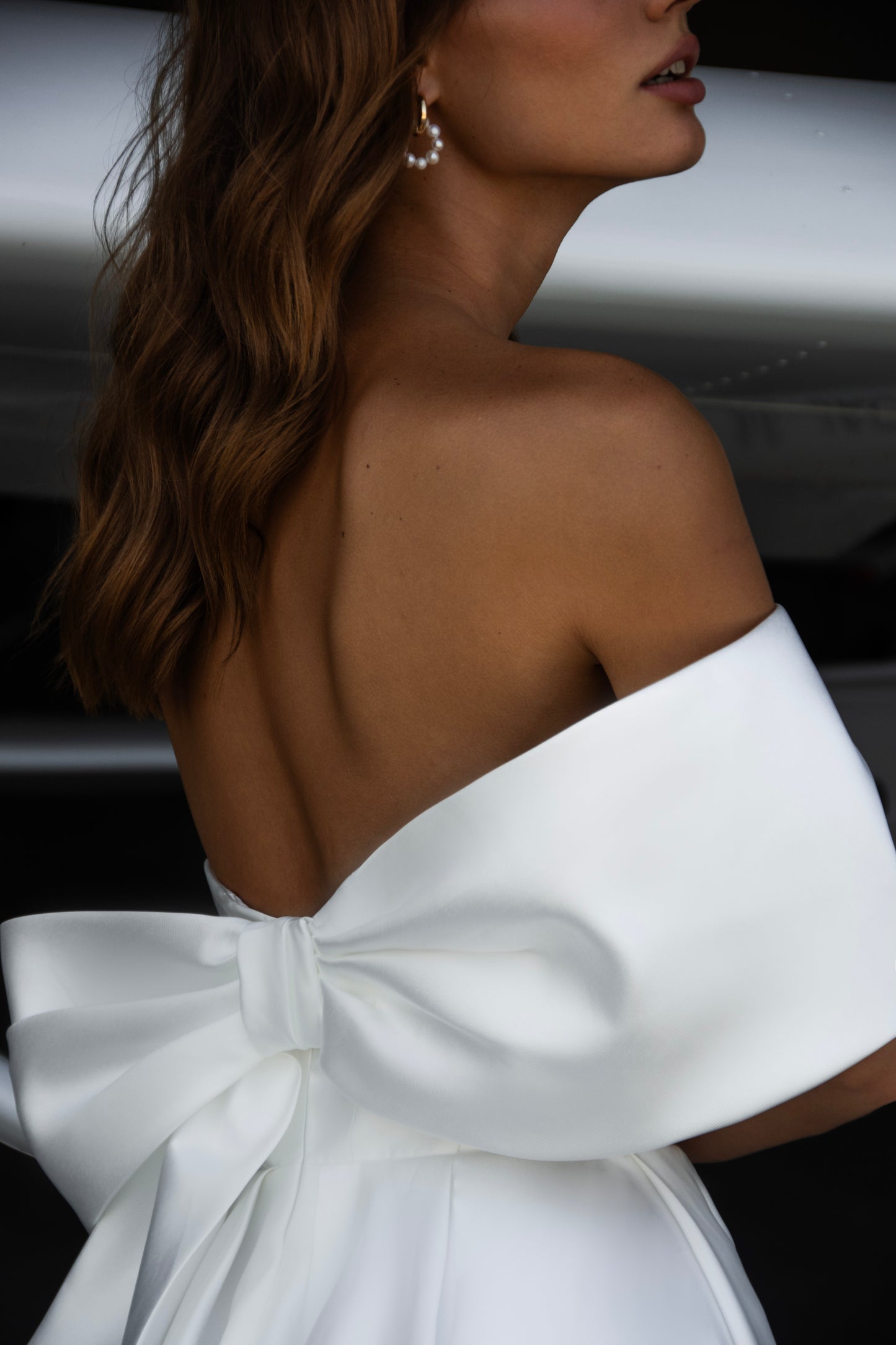 Bride wearing Mikado A-line off-shoulder wedding dress with draped neckline and front slit, standing beside an airplane.
