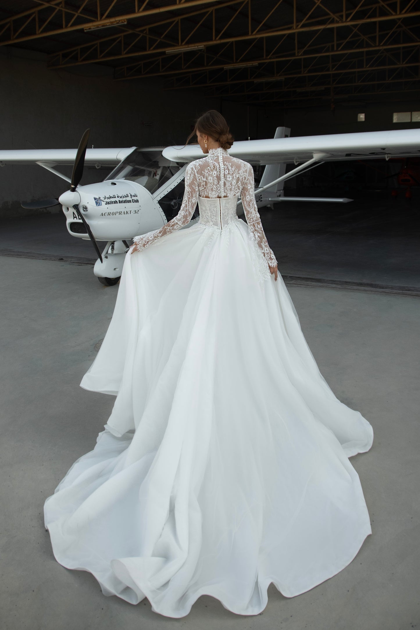 Bride wearing lace long-sleeve wedding dress with high illusion neckline, fitted corset bodice, and chapel train, featuring intricate lace cuffs and back detailing.