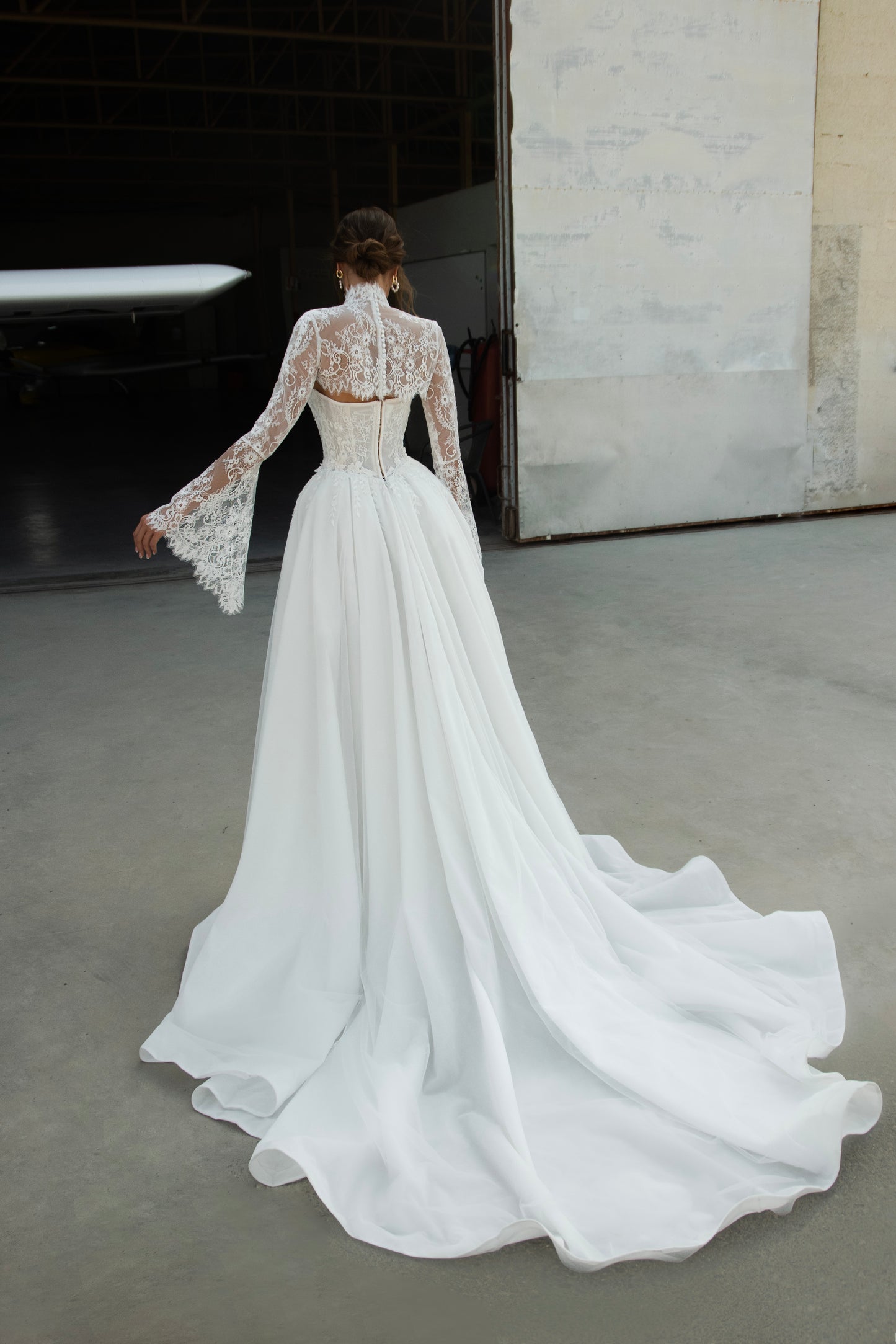 Bride wearing lace long-sleeve wedding dress with high illusion neckline, fitted corset bodice, and chapel train, featuring intricate lace cuffs and back detailing.