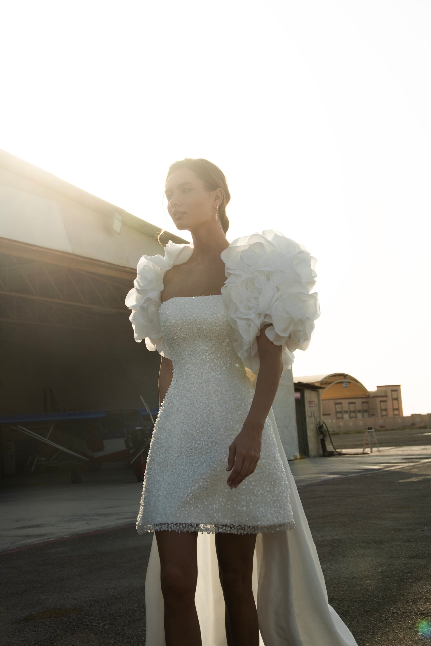 Bride wearing short beaded mini wedding dress with voluminous floral bolero and detachable satin train, photographed at sunset airfield.