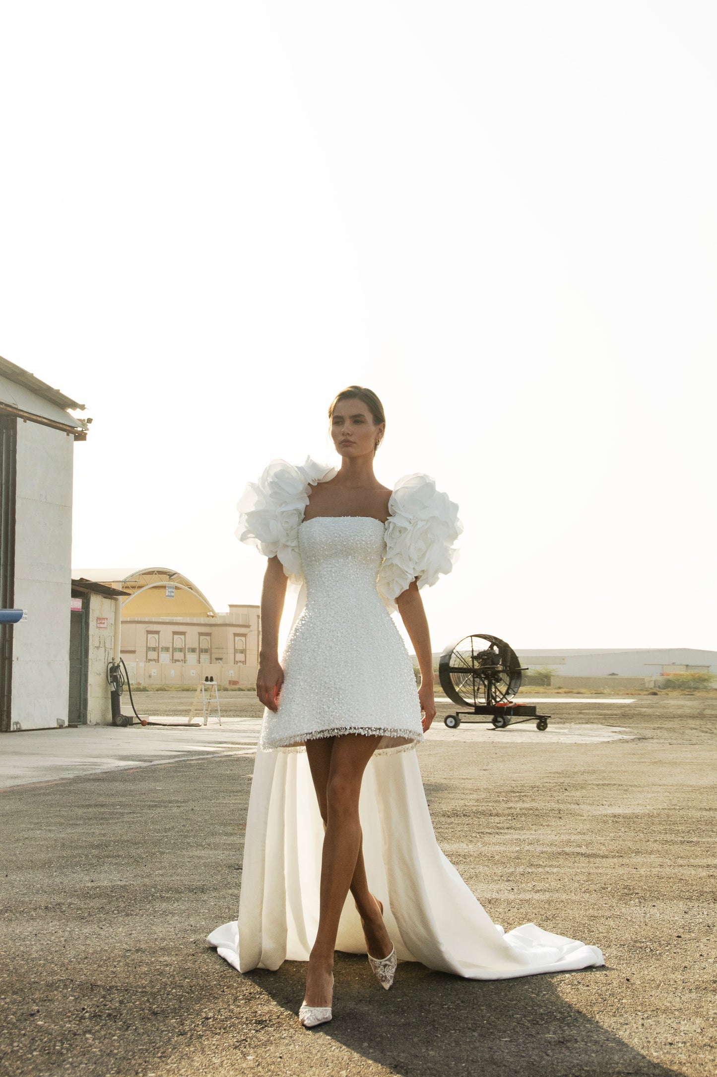 Bride wearing short beaded mini wedding dress with voluminous floral bolero and detachable satin train, photographed at sunset airfield.