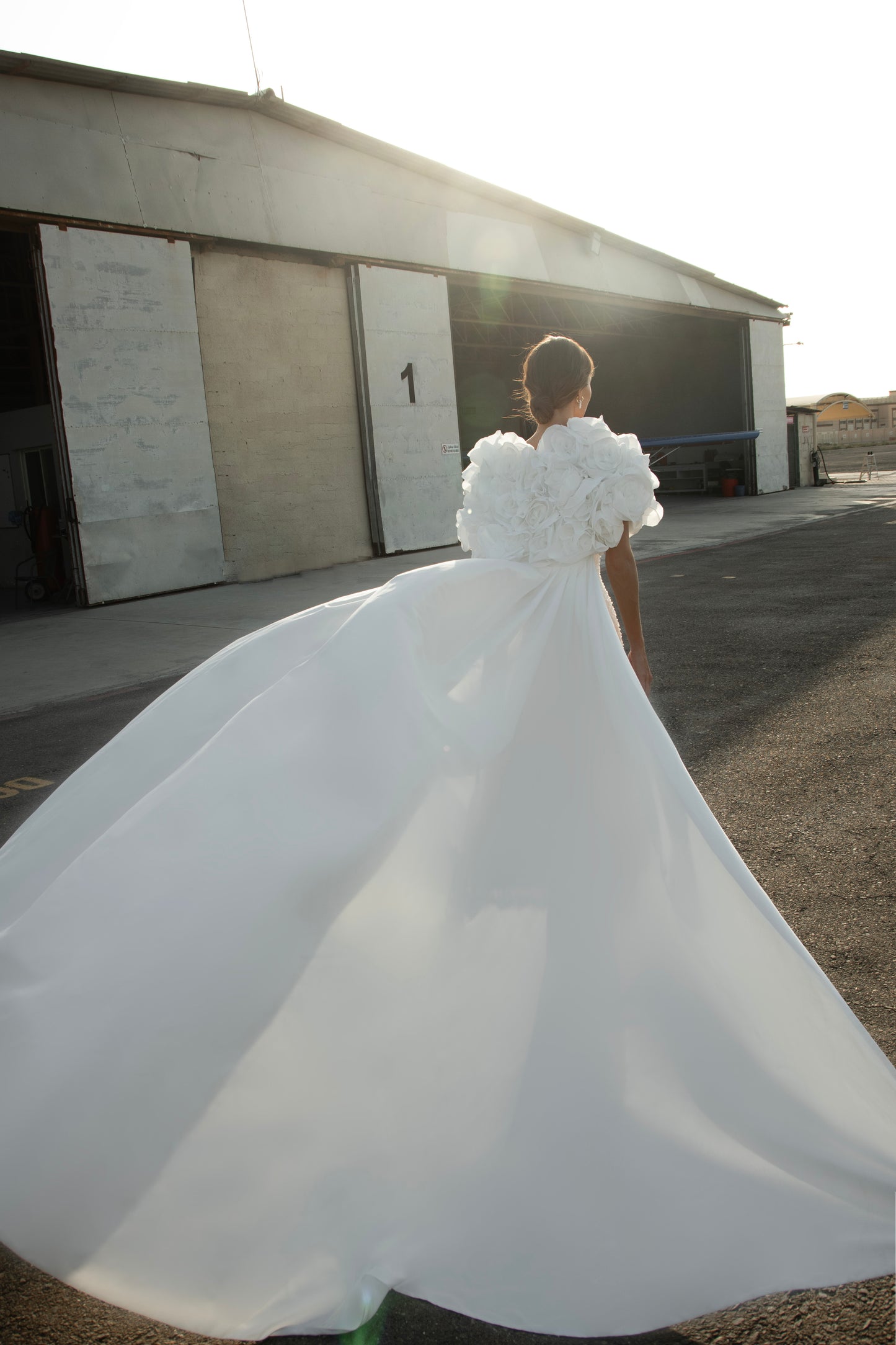 Bride wearing short beaded mini wedding dress with voluminous floral bolero and detachable satin train, photographed at sunset airfield.