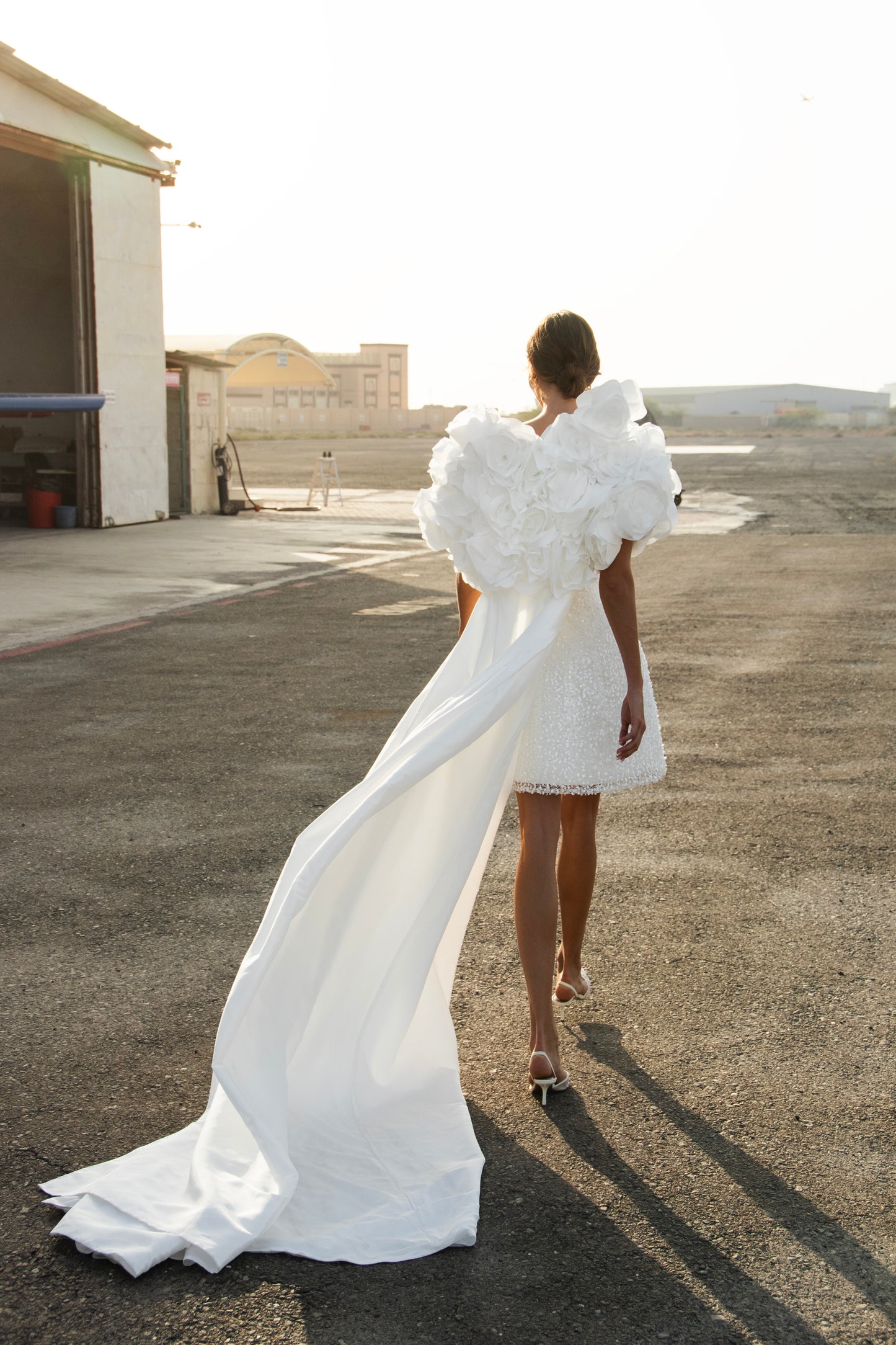 Bride wearing short beaded mini wedding dress with voluminous floral bolero and detachable satin train, photographed at sunset airfield.