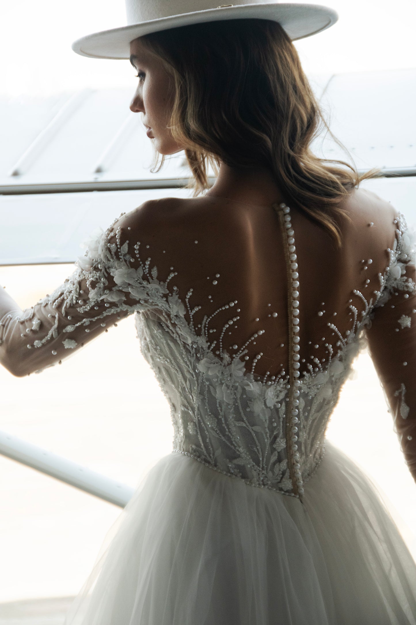 Bride wearing illusion tulle ball gown with 3D floral appliqués, pearl embroidery, and illusion back with buttons, photographed beside an airplane hangar.