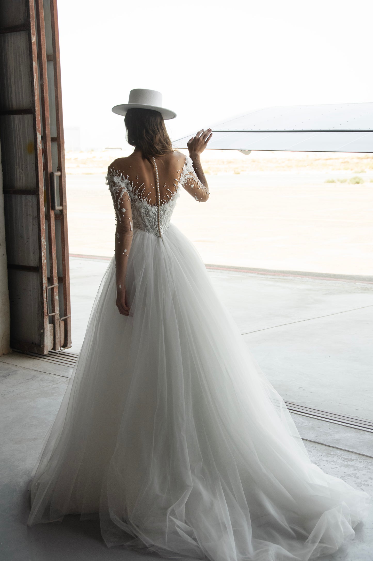 Bride wearing illusion tulle ball gown with 3D floral appliqués, pearl embroidery, and illusion back with buttons, photographed beside an airplane hangar.