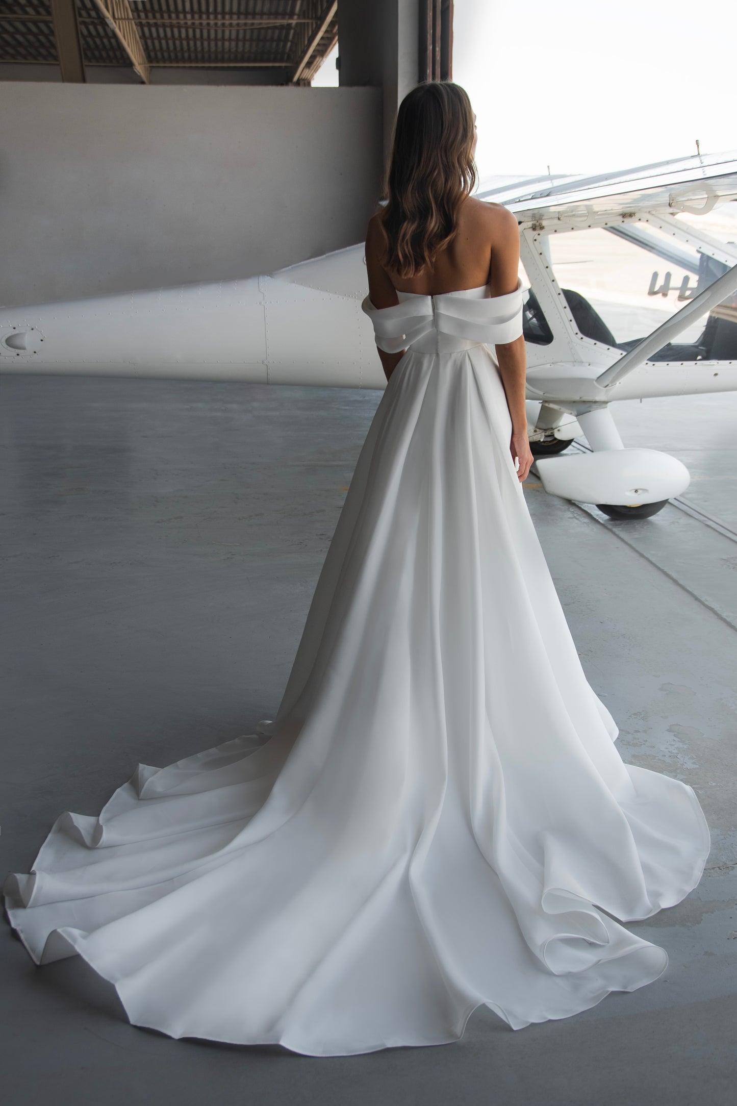 Off-shoulder organza wedding dress with sculpted folded bodice, high-slit skirt, and flowing train in soft white fabric for modern minimalist bridal look