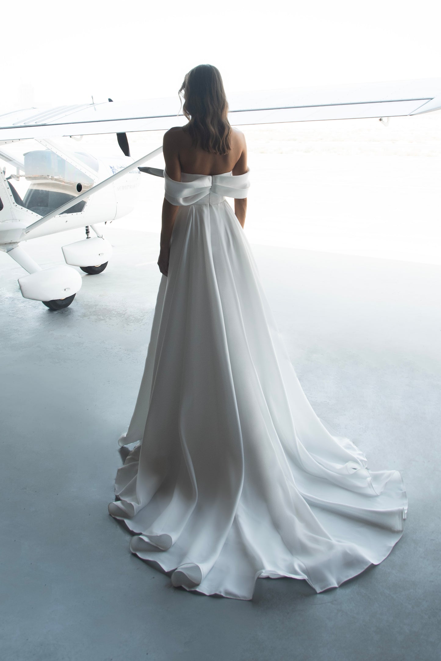 Off-shoulder organza wedding dress with sculpted folded bodice, high-slit skirt, and flowing train in soft white fabric for modern minimalist bridal look