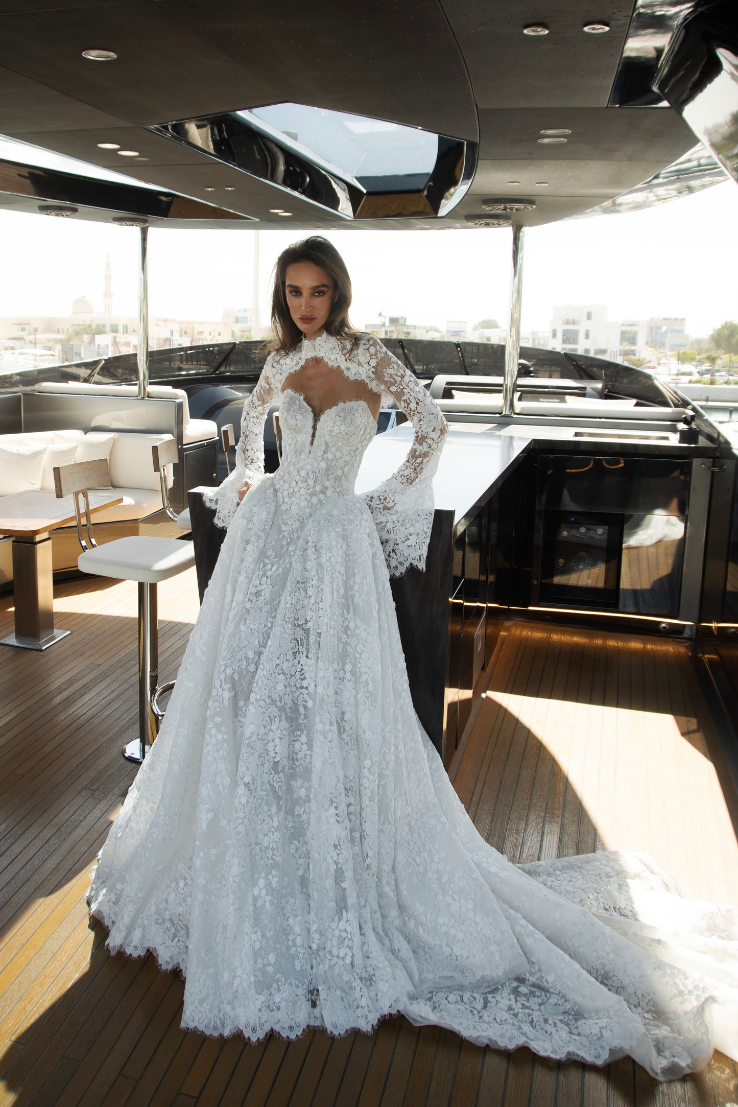 Bride wearing a high-neck lace wedding gown with long bell sleeves and chapel train from the Celeste Collection, photographed on a yacht.