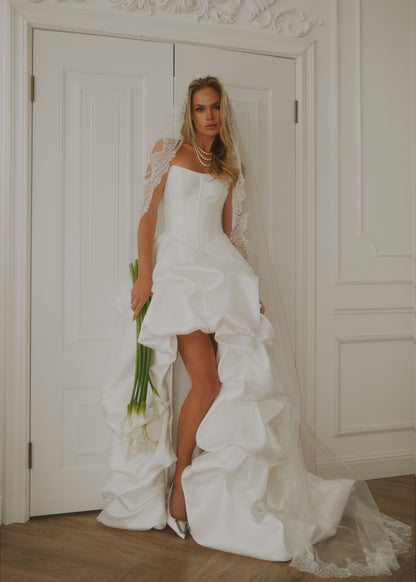 Full-length view of a satin high-low dress with a long ruffled train, in a bright interior
