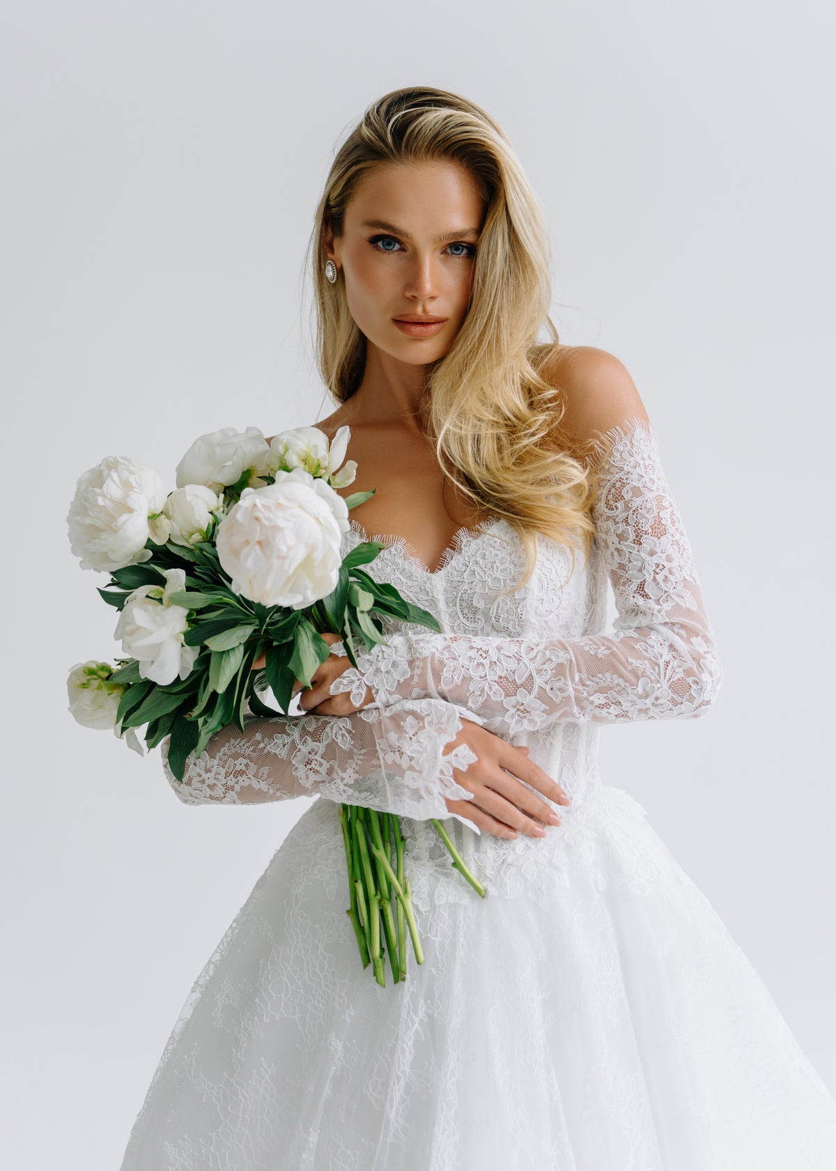 Bride in a lace off-shoulder wedding dress with long sleeves, holding white peonies