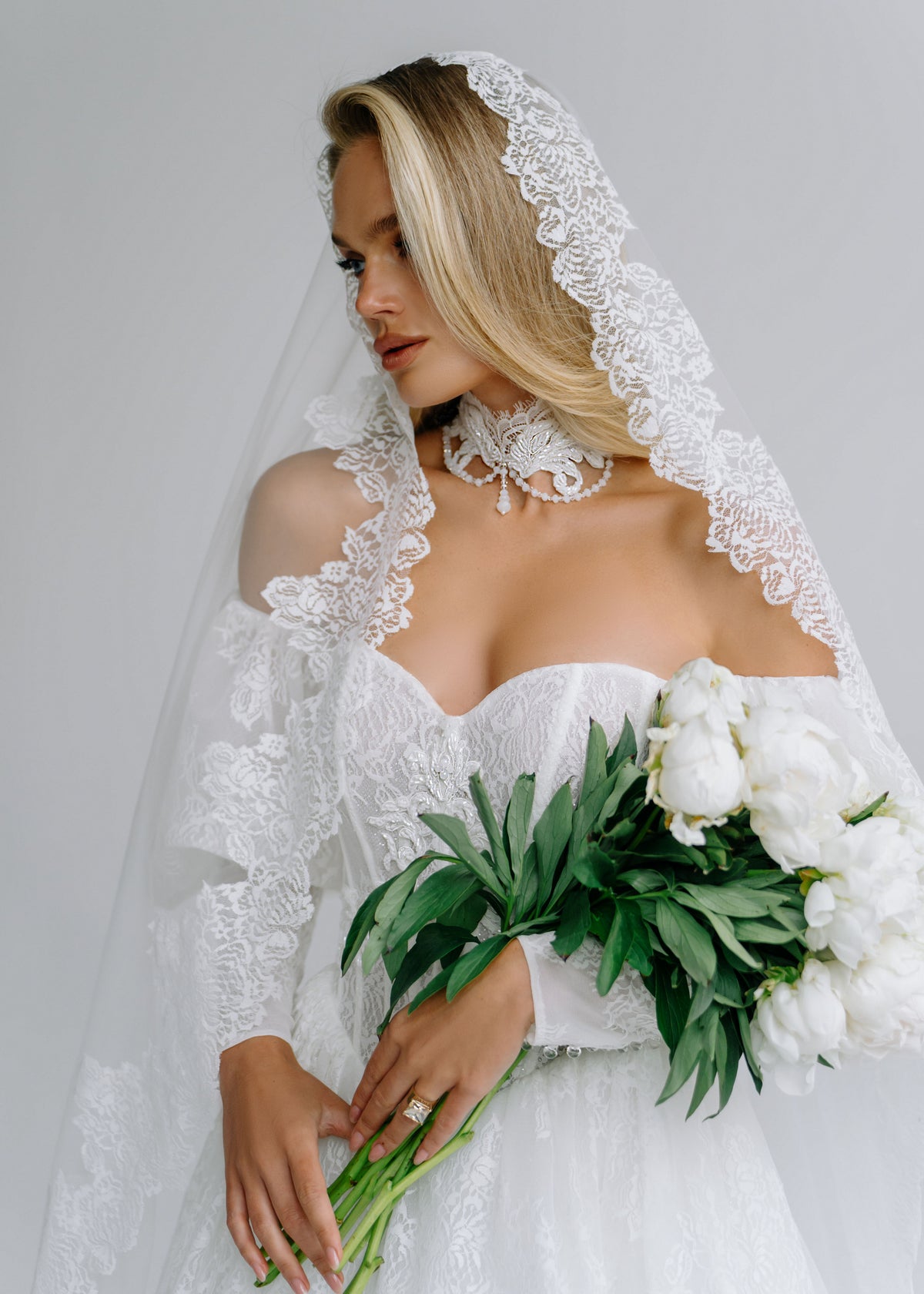 Bride in a lace off-shoulder wedding dress with a scalloped lace-trimmed veil, holding white peonies