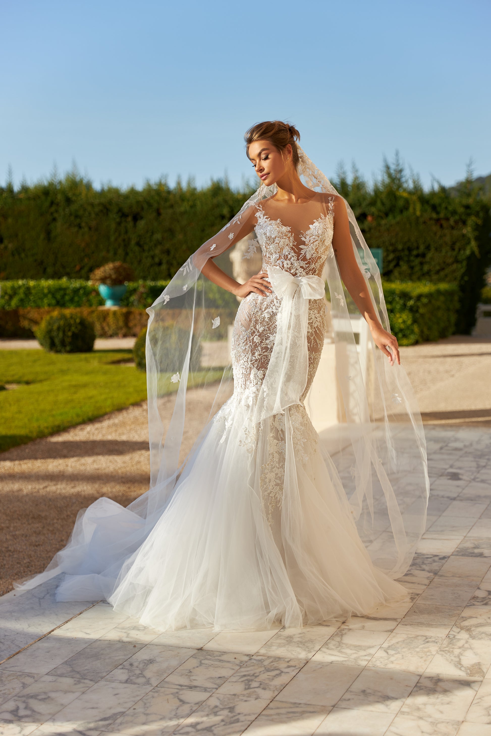 Modern mermaid wedding dress with knee-high cut, detachable waist bow, lace appliques, bead and pearl embroidery, 50 cm train, and matching veil. Made of lace and tulle for a timeless bridal look.