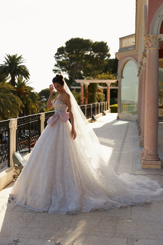 A-line bridal gown with sleeveless bodice, open back, blush taffeta belt and bow, chapel train, and lace appliques with pearl and sequin embroidery. Made from Chantilly lace.