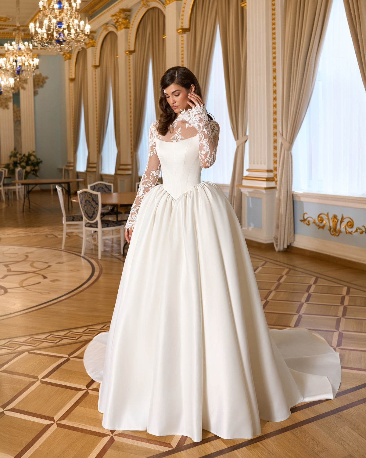Full-length front view of an A-line wedding dress with a pointed basque waist, floral lace bodice layer and long sheer sleeves.