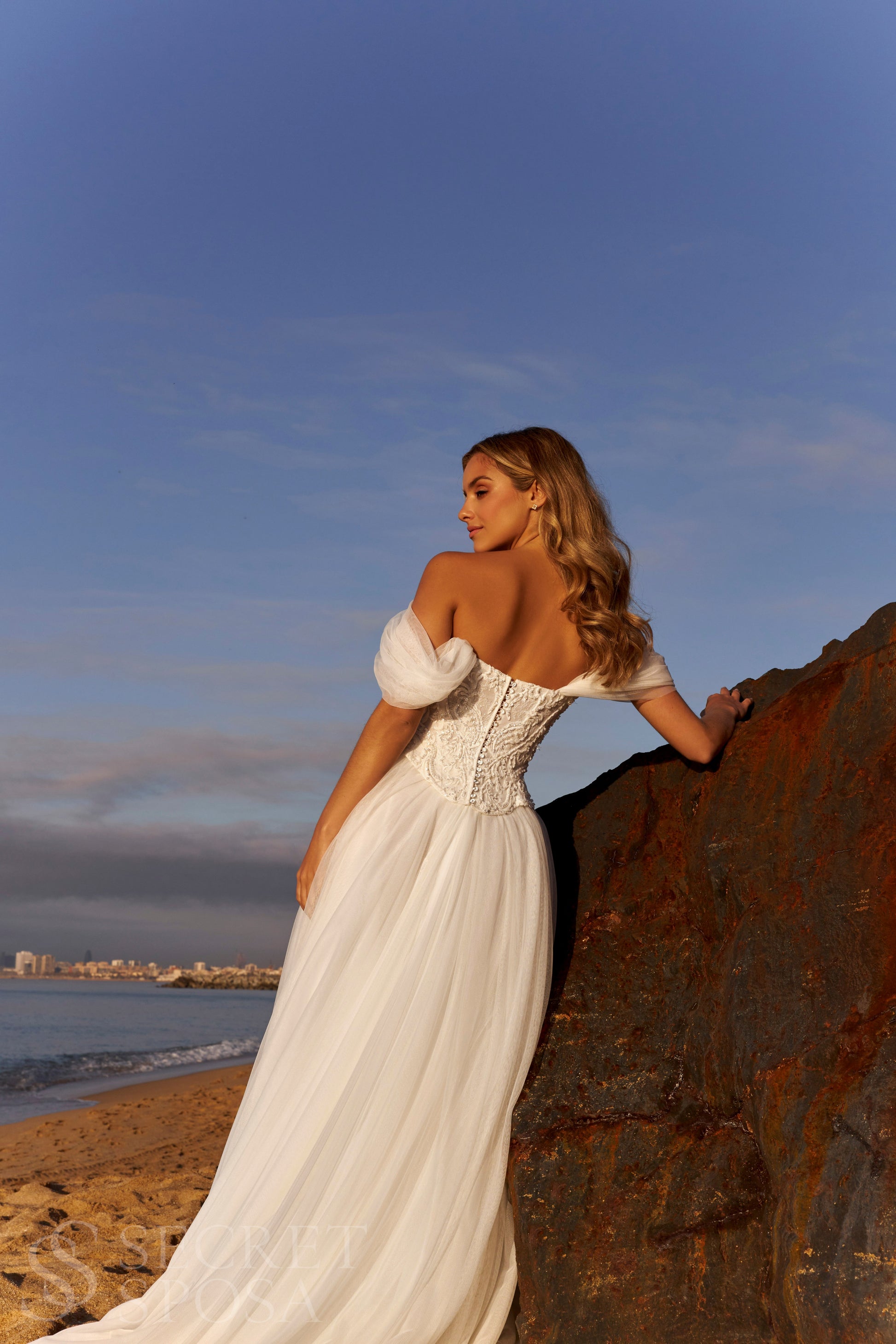 Bride in romantic A-line gown with off-the-shoulder draped tulle sleeves, lace corset bodice, V-neckline, and slit tulle skirt.