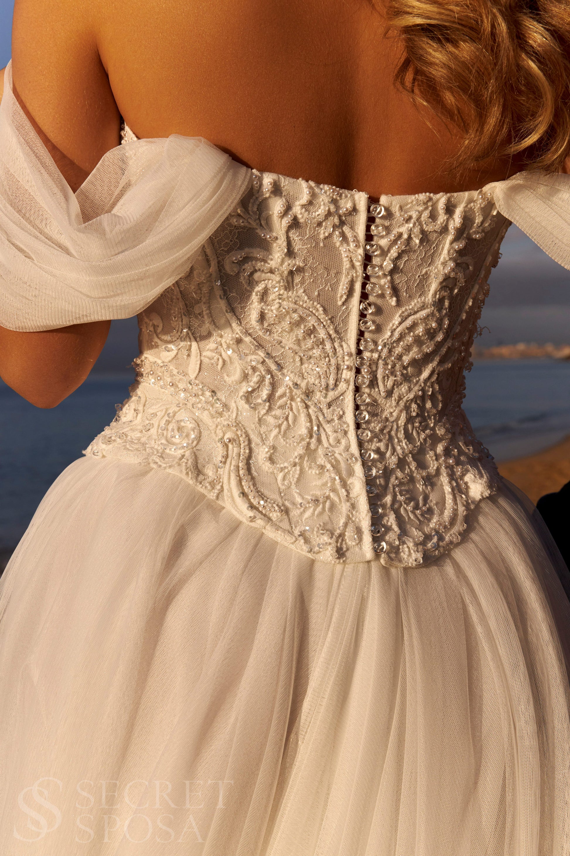 Bride in romantic A-line gown with off-the-shoulder draped tulle sleeves, lace corset bodice, V-neckline, and slit tulle skirt.