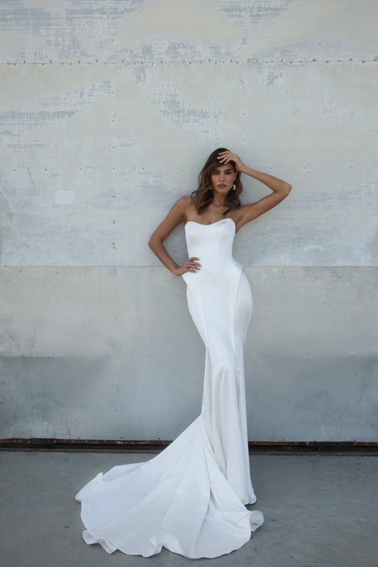Bride wearing strapless satin mermaid wedding dress with structured sides and chapel train, photographed in a hangar beside an airplane.