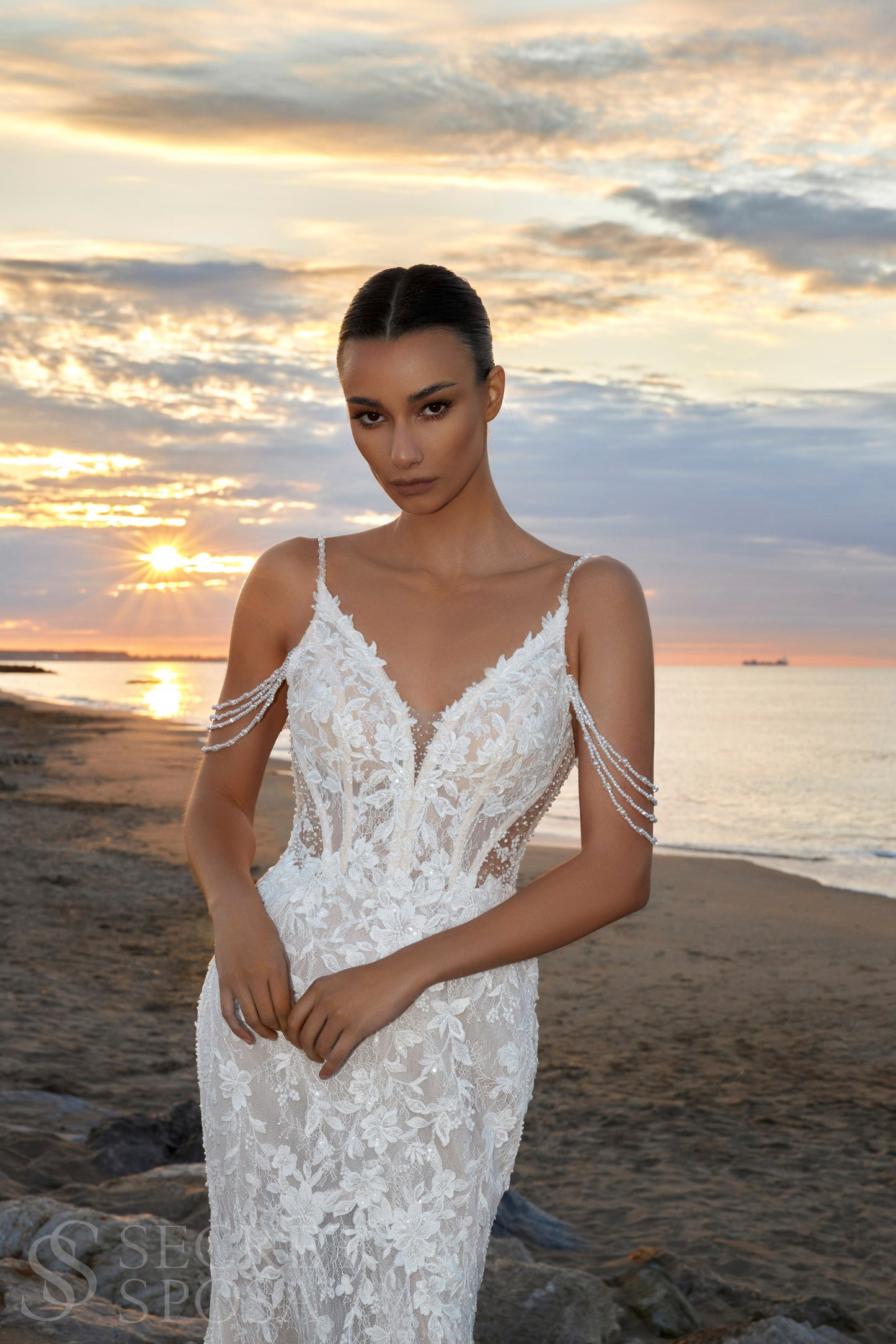 Bride in mermaid fit-and-flare lace wedding dress with pearl spaghetti straps, off-the-shoulder pearl strands, V-neckline, and sweep train.