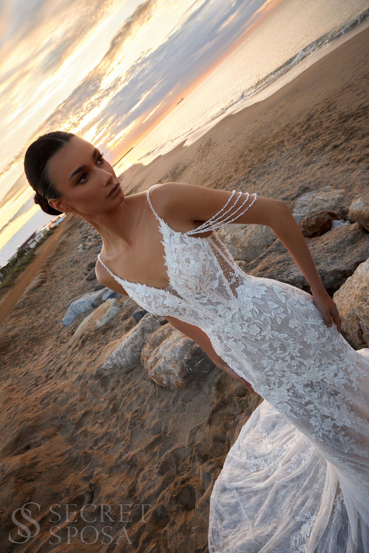 Bride in mermaid fit-and-flare lace wedding dress with pearl spaghetti straps, off-the-shoulder pearl strands, V-neckline, and sweep train.