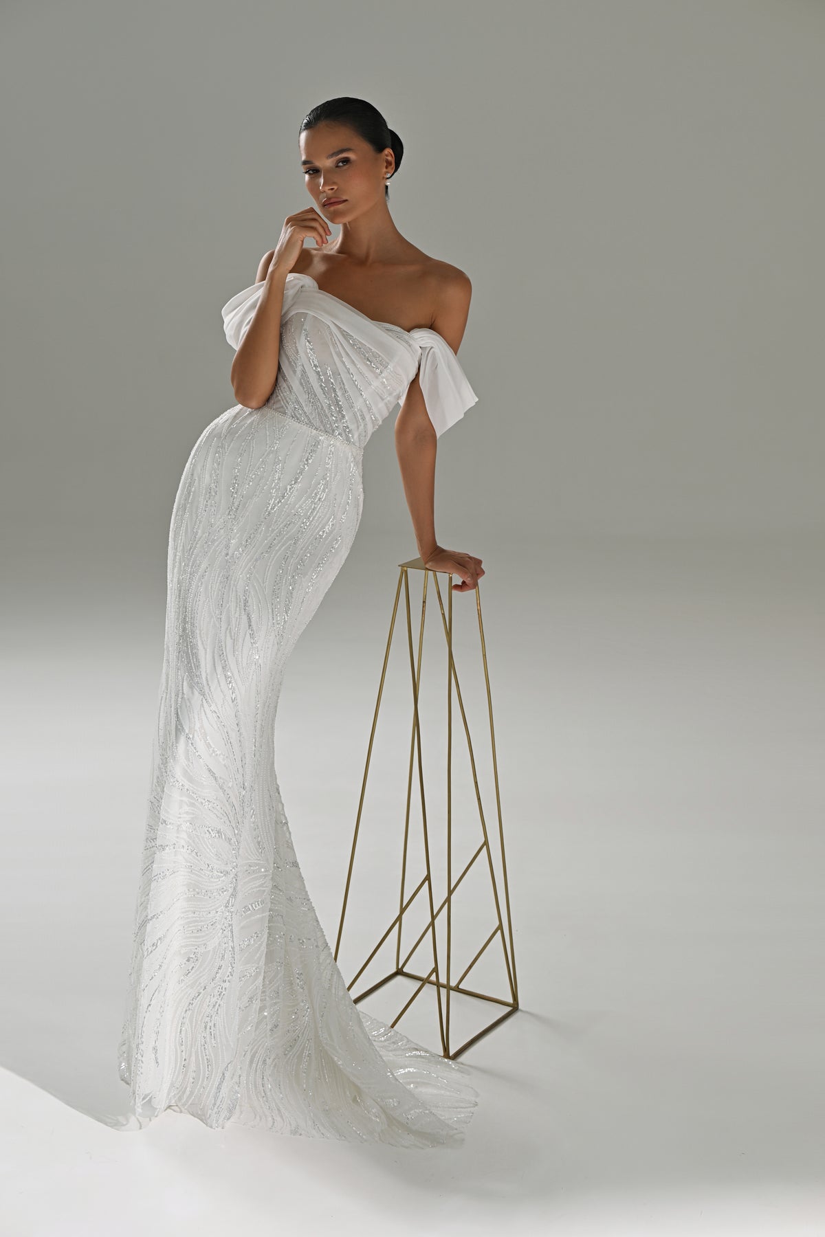 Lace mermaid wedding dress with asymmetric off-shoulder drape – model leaning on a gold stand, full-length view