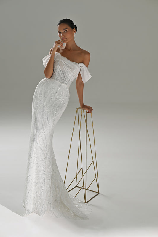 Lace mermaid wedding dress with asymmetric off-shoulder drape – model leaning on a gold stand, full-length view